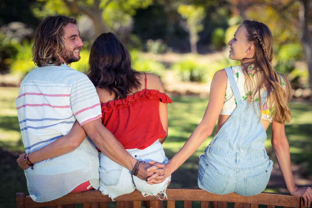Three young adults sitting on a bench outdoors, two holding hands behind the back of the middle person.