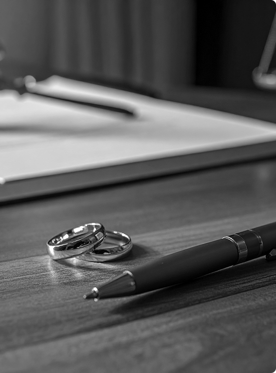 Two silver wedding bands beside a black pen on a wooden desk with blurred documents in the background.