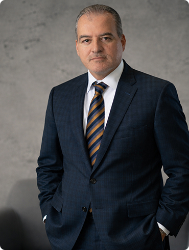 Middle-aged man in a navy suit and striped tie standing confidently against a grey textured background.